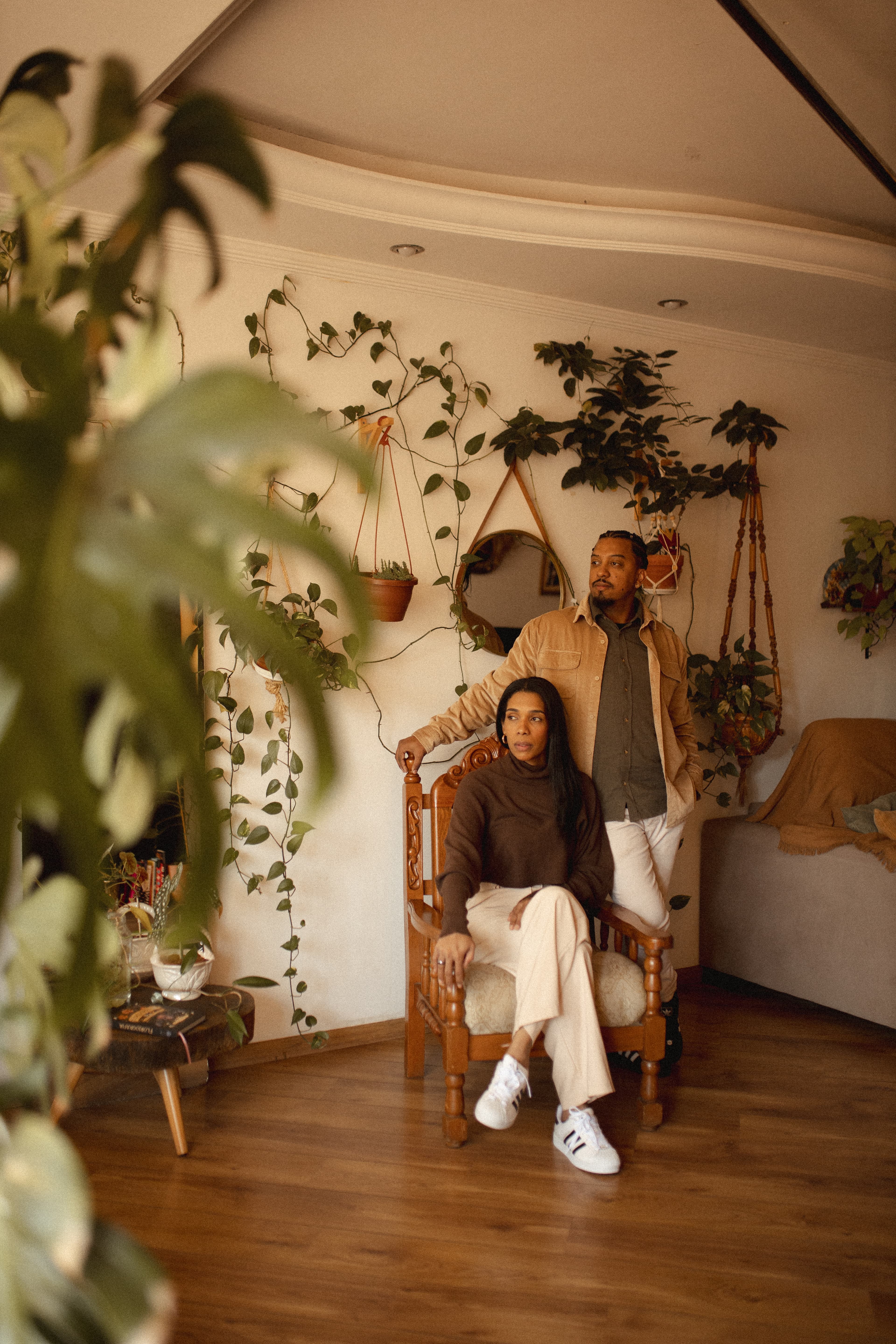 Paula sentada em uma cadeira de madeira, com Erik ao seu lado, entre as plantas da sala.